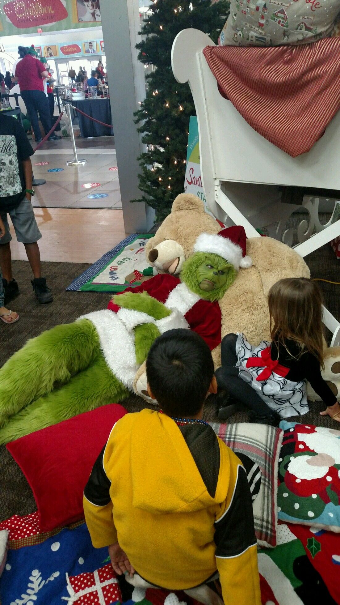 Costumed Grinch rests on overstuffed teddy bear and talks to young children.