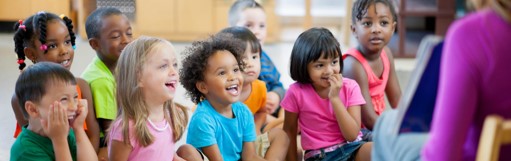 happy children sitting down on classroom carpet smiling while looking at a teacer